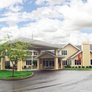 A large, modern building with a covered entrance and surrounding greenery under a partly cloudy sky.