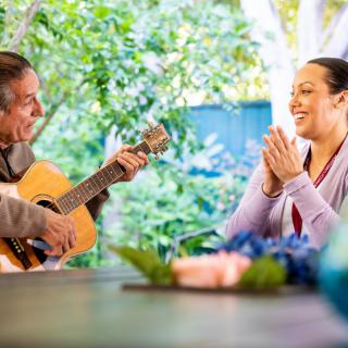 A person playing guitar while another enjoys and smiles in a serene outdoor setting.