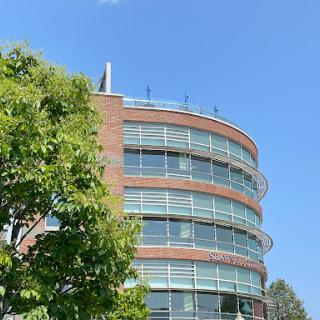 A modern curved building featuring glass windows, surrounded by greenery under a clear blue sky.
