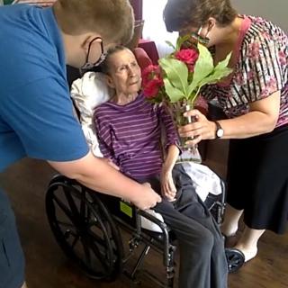 An elderly person in a wheelchair receives flowers from two caregivers.