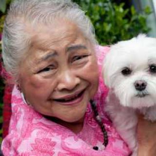 An elderly woman joyfully holding her small white dog outside.