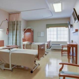 A clean and well-lit hospital room with two patient beds, chairs, and a window.