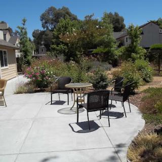 A sunny outdoor patio area with chairs and a small table, surrounded by lush greenery.