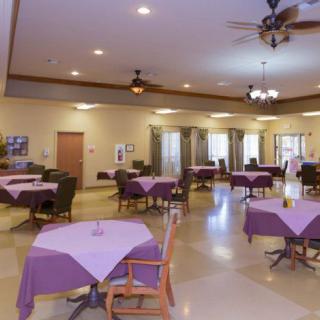 A spacious dining room with tables covered in purple tablecloths, surrounded by chairs and illuminated by ceiling lights and fans.