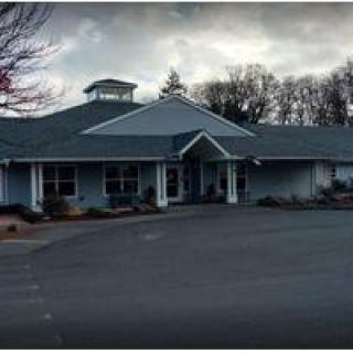 A single-story building with a gabled roof, surrounded by trees and set beside a parking lot.