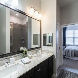 A modern bathroom with a double sink and large mirror, showcasing sleek interior design and a glimpse into an adjoining bedroom.