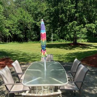 A sunny patio with a glass-top table, surrounded by six chairs and a colorful umbrella, set in a garden with green grass and trees.