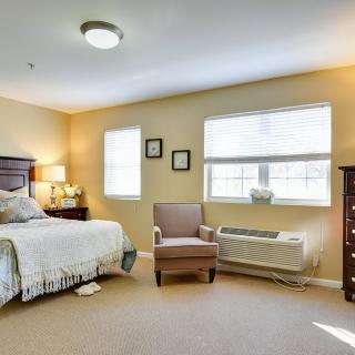 A cozy bedroom featuring a bed, armchair, and dresser with natural light streaming through the windows.