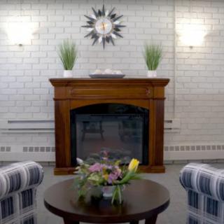 A cozy living room featuring a wooden fireplace with decorative plants, surrounded by striped chairs and a round coffee table with a floral arrangement.