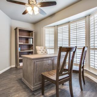 A cozy home office with a wooden desk and chairs, featuring large windows with shutters.