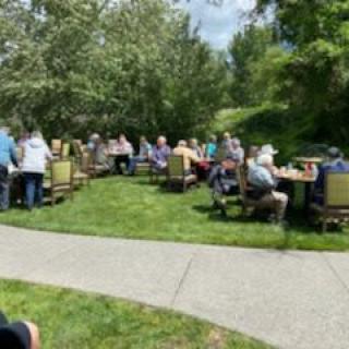A group of people enjoying an outdoor gathering with tables and chairs arranged on a grassy area under sunny skies.