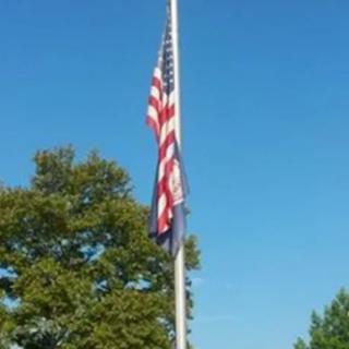An American flag on a flagpole, set against a clear blue sky with trees in the background.