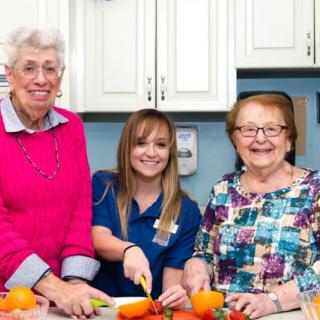 A caregiver and two elderly women are smiling while preparing vegetables in a kitchen.