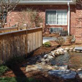 A backyard garden featuring a small pond, surrounded by a wooden fence and adjacent to a brick house.