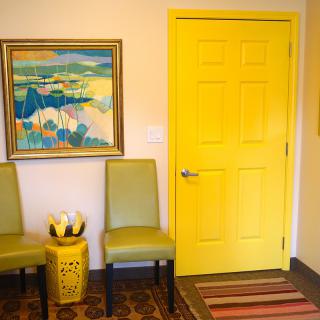 A vibrant room featuring a bright yellow door, green chairs, and a colorful painting on the wall.