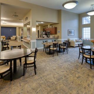 A cozy dining area with multiple tables and chairs, well-lit with ceiling lights and natural light from large windows.
