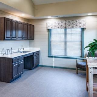 A modern kitchen space with dark cabinets, a double sink, a window with floral curtains, and a small seating area.