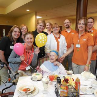 A cheerful group of people celebrating with an elderly person at a dining table, decorated with colorful balloons.