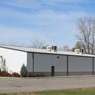 An exterior view of a simple industrial warehouse with a parking lot and trees in the background.