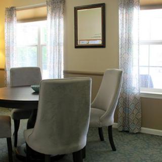 A cozy dining room with upholstered chairs and a round table, flanked by large windows draped in sheer curtains.