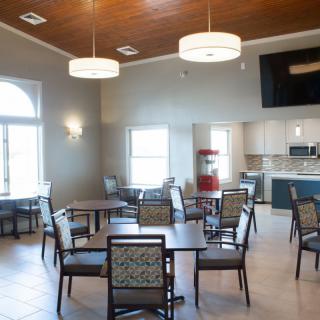 A modern community dining area with several tables and chairs, featuring a kitchen in the background.