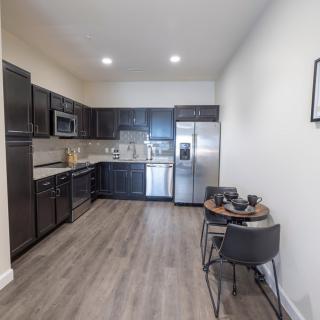 A modern kitchen with dark cabinetry and stainless steel appliances, featuring a small dining area with two chairs on the side.