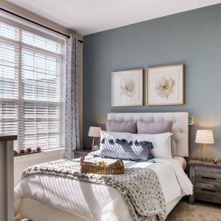 A cozy bedroom with stylish decor, featuring a bed, artwork on the wall, and ample natural light from a large window.