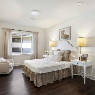 A cozy bedroom with modern white decor, featuring a bed, lamps, and a comfortable chair by the window.