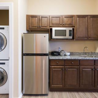 A compact kitchen and laundry area featuring wooden cabinets, a stainless steel refrigerator, microwave, and stacked washer and dryer.