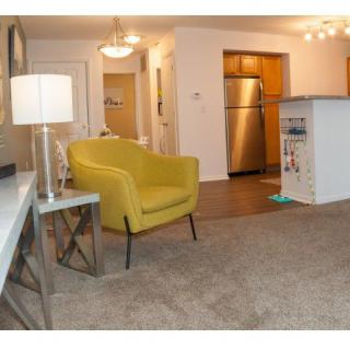 A modern open-plan living area featuring a yellow armchair, a lamp, and a view of the kitchen with stainless steel appliances.