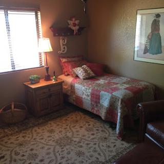 A cozy bedroom featuring a single bed dressed in a colorful quilt, accompanied by a nightstand and lamp, with natural light flowing through a window.