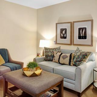 A cozy living room with a sofa, armchair, and coffee table, accessorized with cushions and artwork on the wall.