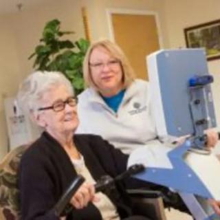 An elderly woman using exercise equipment with the assistance of a caregiver in a healthcare setting.