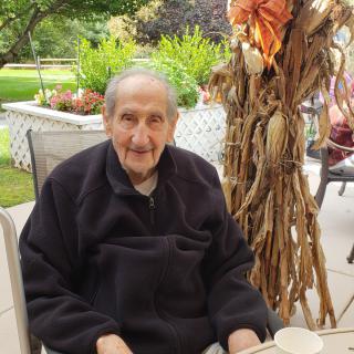 An elderly man sits outdoors, smiling with autumn decorations nearby.