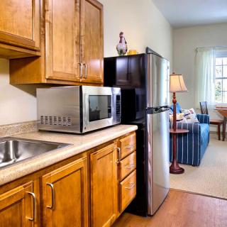 A compact kitchen area with wooden cabinets featuring a microwave, refrigerator, and sink, adjacent to a cozy living room.