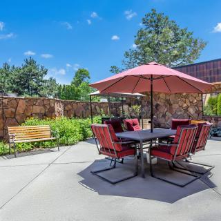 A sunny patio with a table and chairs set under a large red umbrella, surrounded by a garden.