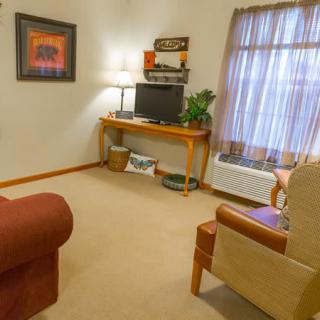 A cozy living room with a sofa, television on a wooden desk, and a lamp beside framed artwork.
