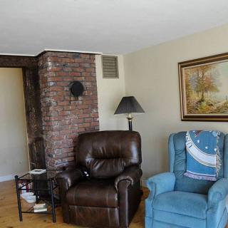 A cozy living room with a brick wall, featuring a brown leather armchair and a blue armchair next to a lamp and painting.