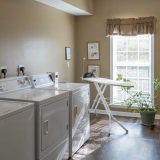 A well-lit laundry room with a washing machine, dryer, ironing board, and a potted plant by the window.