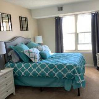 A cozy bedroom with a teal and white bedspread, multiple pillows, and two windows with dark curtains.