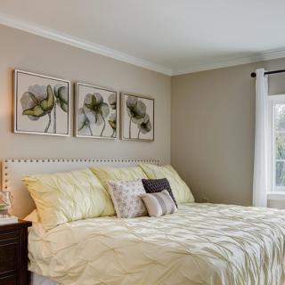A cozy bedroom with a neatly made bed, featuring floral artwork above the headboard and soft natural lighting from a nearby window.