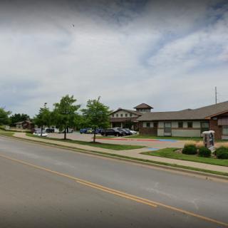 A street view showing a large building with a parking lot, surrounded by trees and a sidewalk under a cloudy sky.