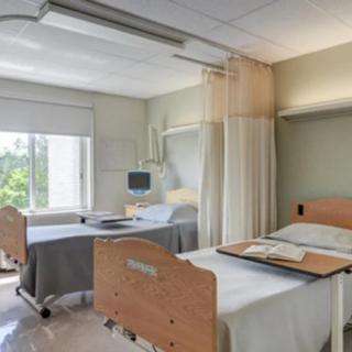 An empty hospital room with two neatly made beds, separated by a curtain, and equipped with a monitor near the window letting in natural light.