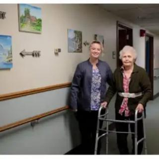 An elderly woman with a walker stands in a hallway with another person next to her, surrounded by wall-mounted paintings.