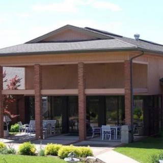 A modern building with a shaded porch featuring tables and chairs, surrounded by a well-maintained garden under a sunny sky.