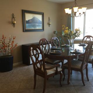 A well-lit dining room with a glass table, wooden chairs, and decorative plants.