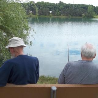 Two elderly men sitting on a bench, fishing by a tranquil lake.