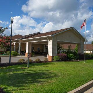 A building entrance with a covered driveway and an American flag flying, surrounded by well-maintained landscaping under a partly cloudy sky.
