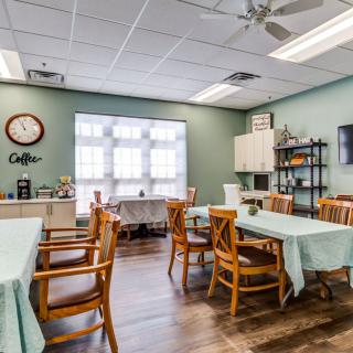 A cozy dining area with tables and chairs, decorated with a clock and natural light from large windows.