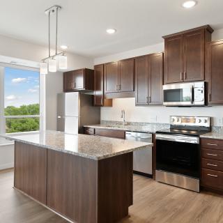 A modern kitchen with wooden cabinets and stainless steel appliances, featuring a central island and large window.